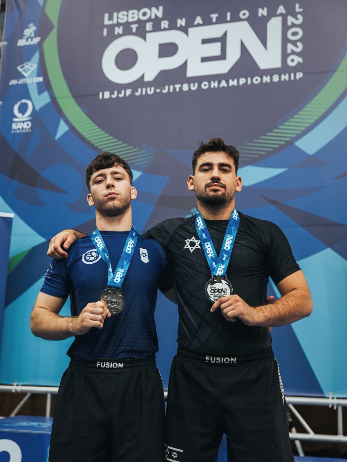 שלו אלנברג ולב טוקר מ-Fusion BJJ חיפה עם מדליות הכסף מול שלט Lisbon International Open של IBJJF